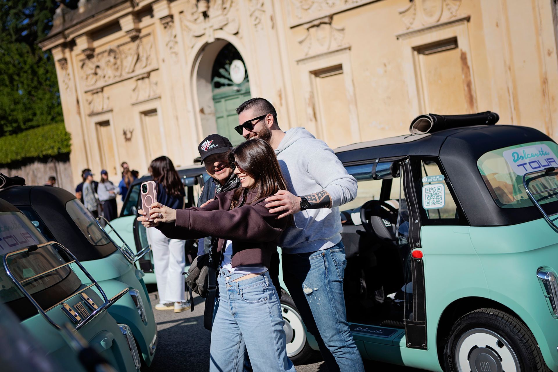 Two people taking a selfie beside a mint-green car in front of a historic building