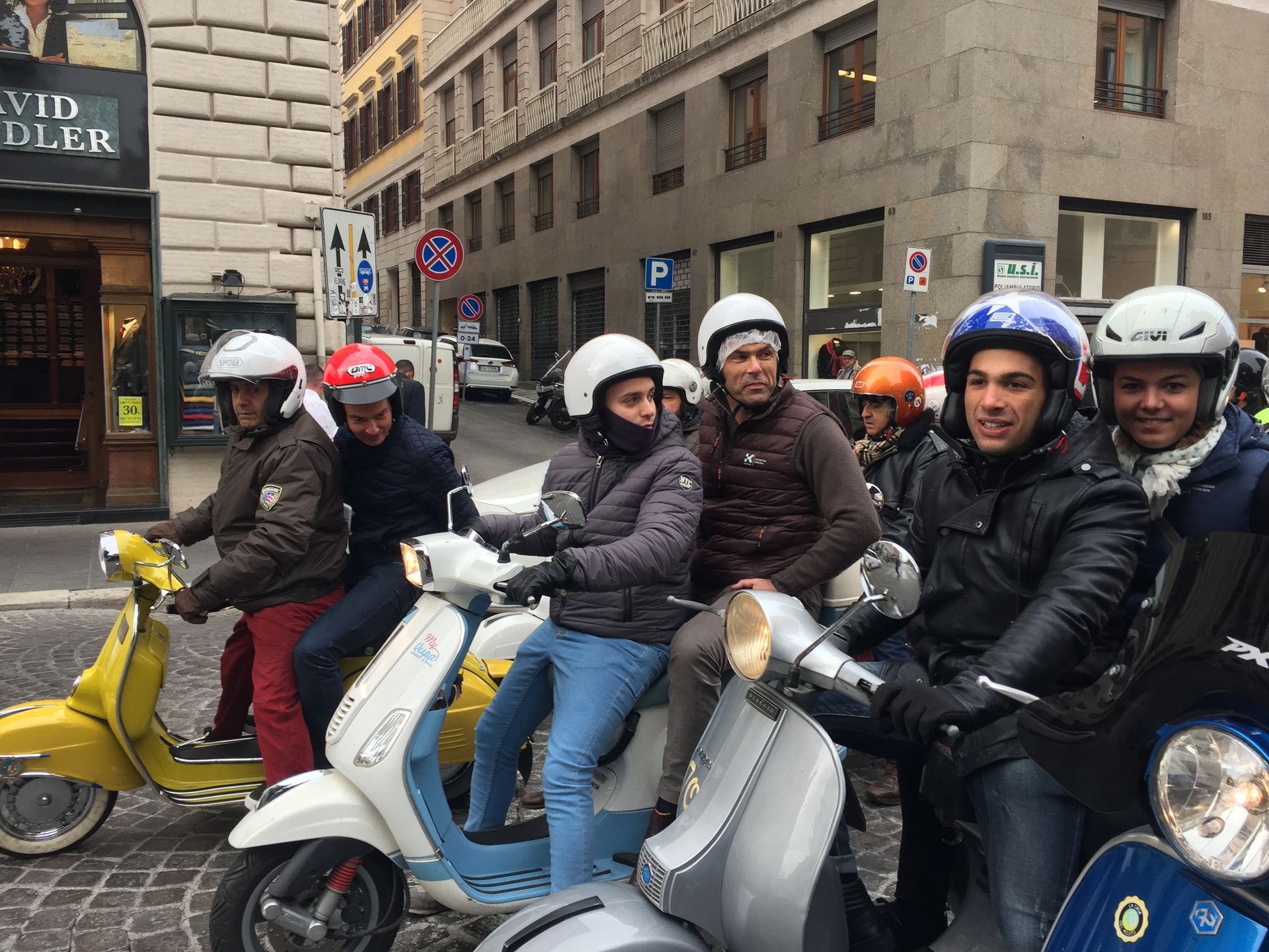 Five helmeted riders on scooters stopped on a city street, with one yellow scooter in front