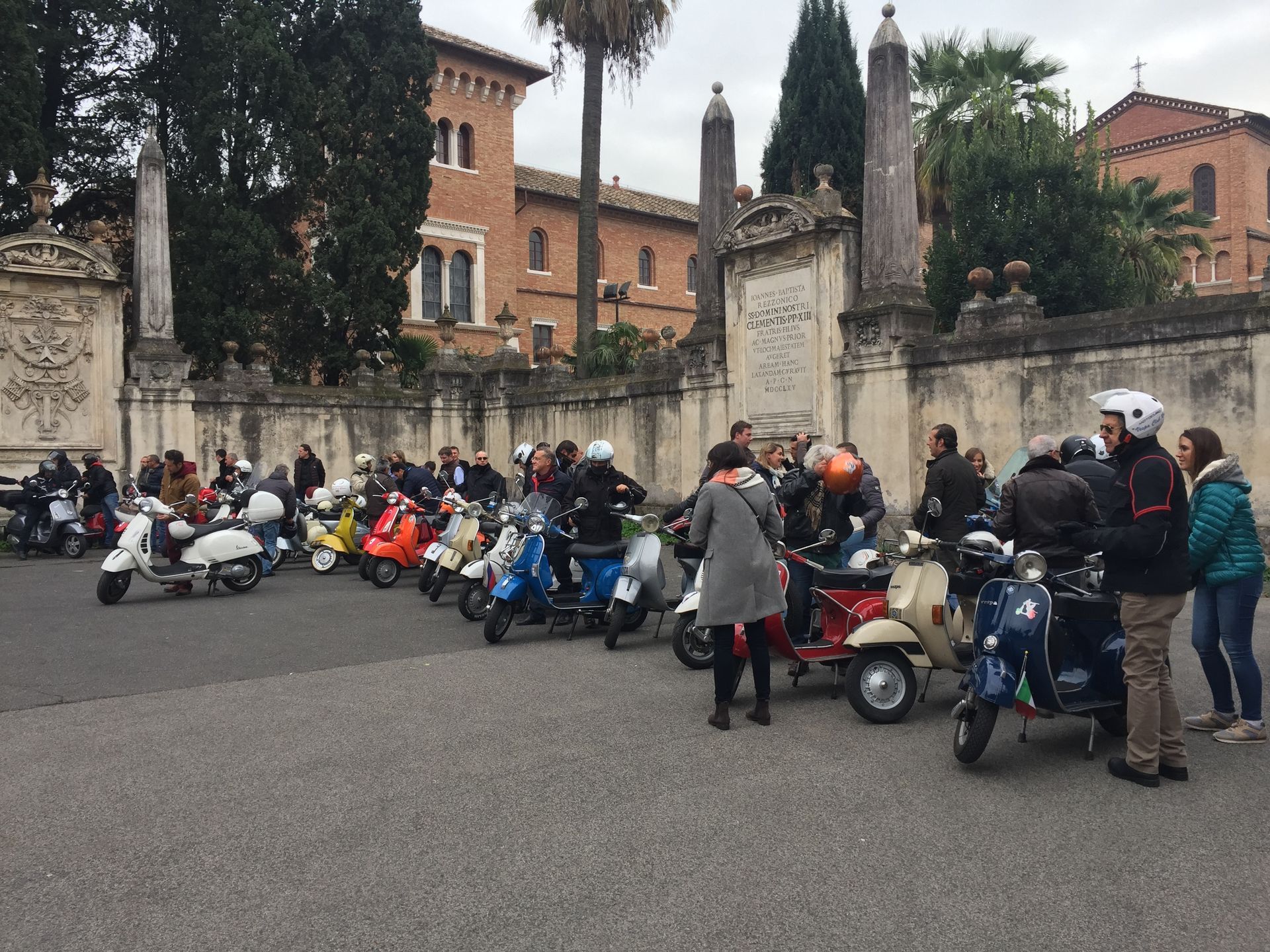 Scooters lined up by a stone wall near a historic building, with riders gathered in a courtyard.