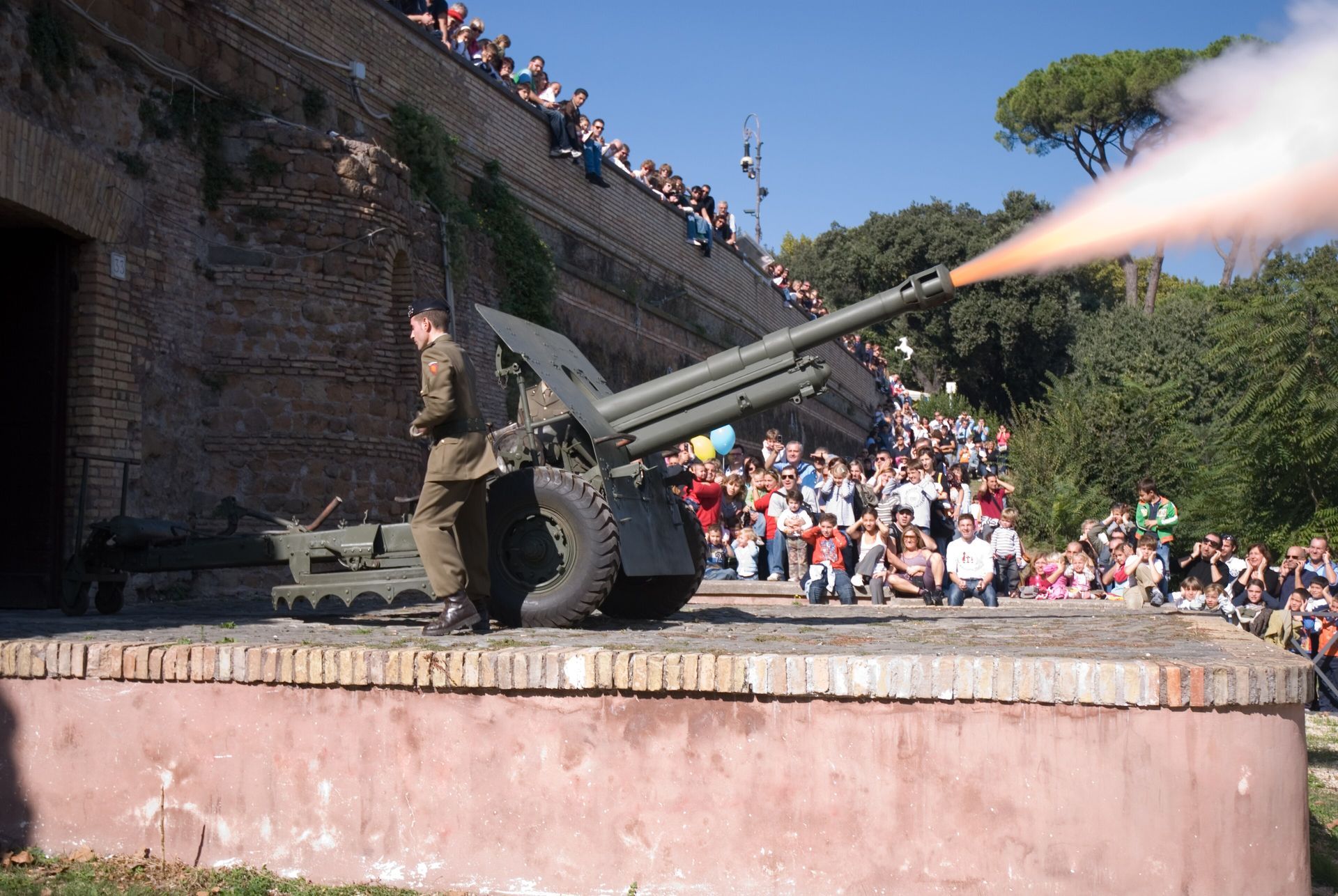 Cannon firing at a stone fort, with a crowd watching from the walls and courtyard.