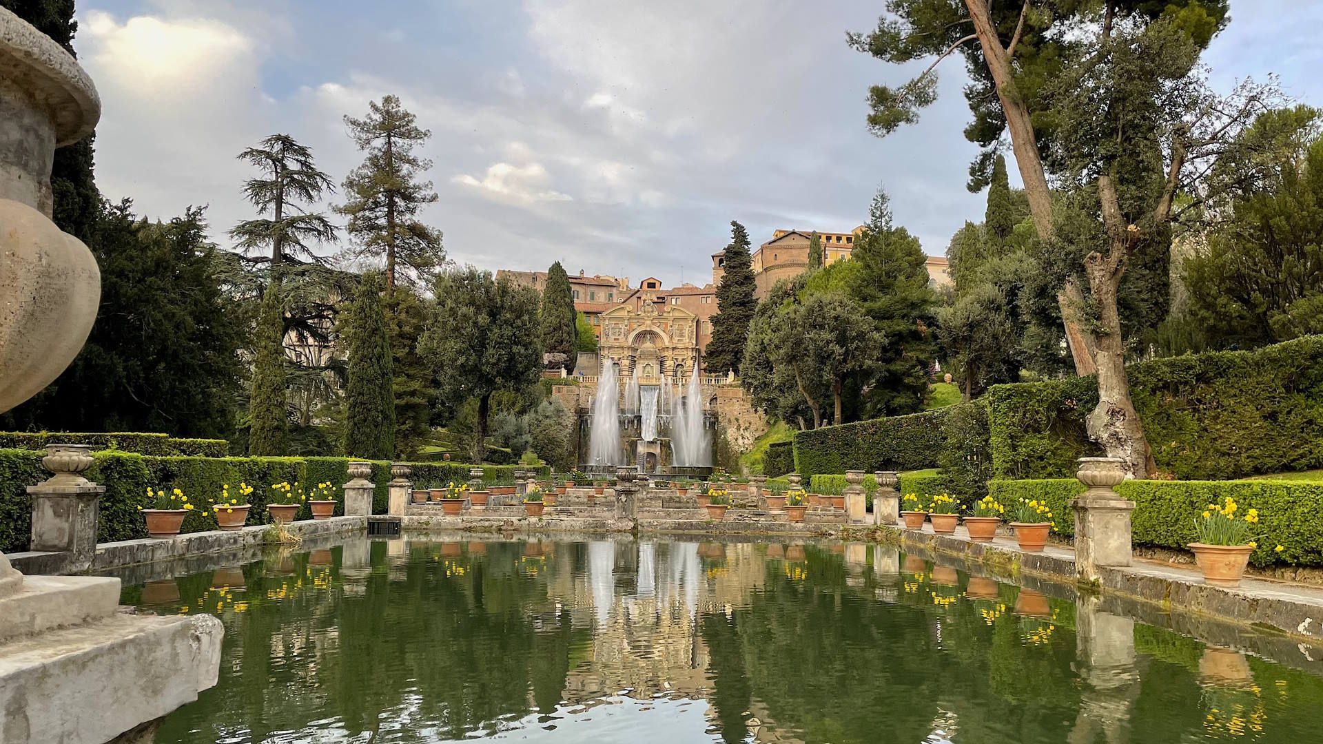 Formal garden with reflecting pool, fountains, and a building framed by trees and statues