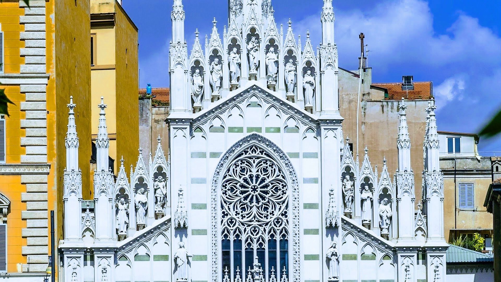 White Gothic church facade with ornate spires and a large rose window among colorful buildings under a blue sky