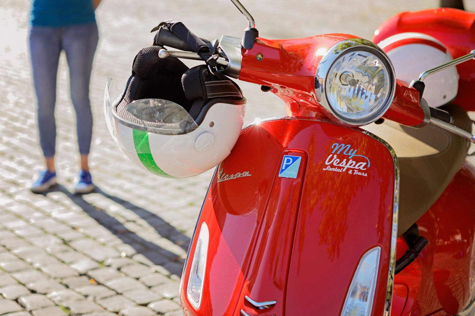 Red scooter with a white helmet hanging on the handlebar, parked on a cobblestone street.
