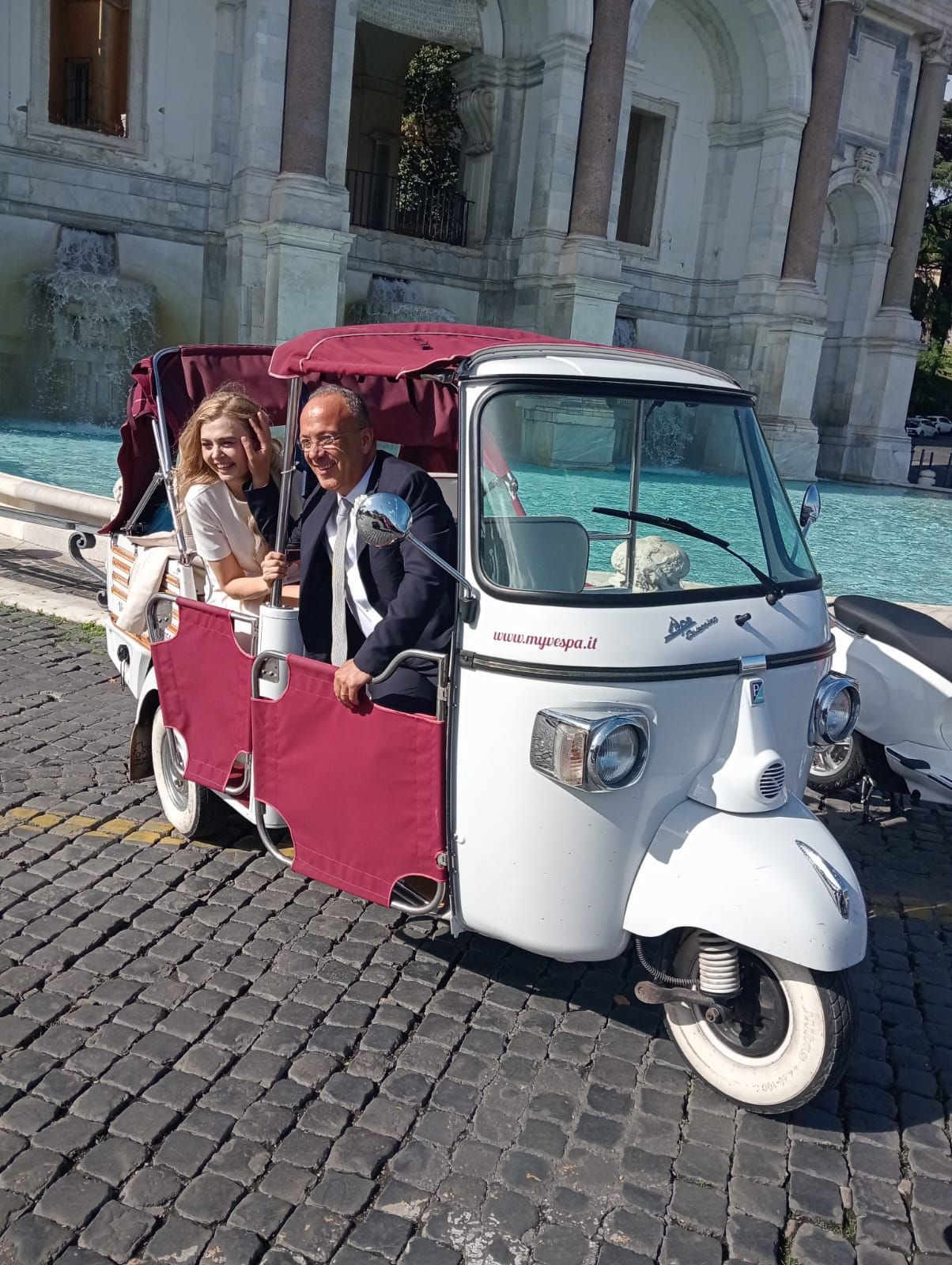 Two people riding in a red-and-white tuk-tuk on a cobblestone street