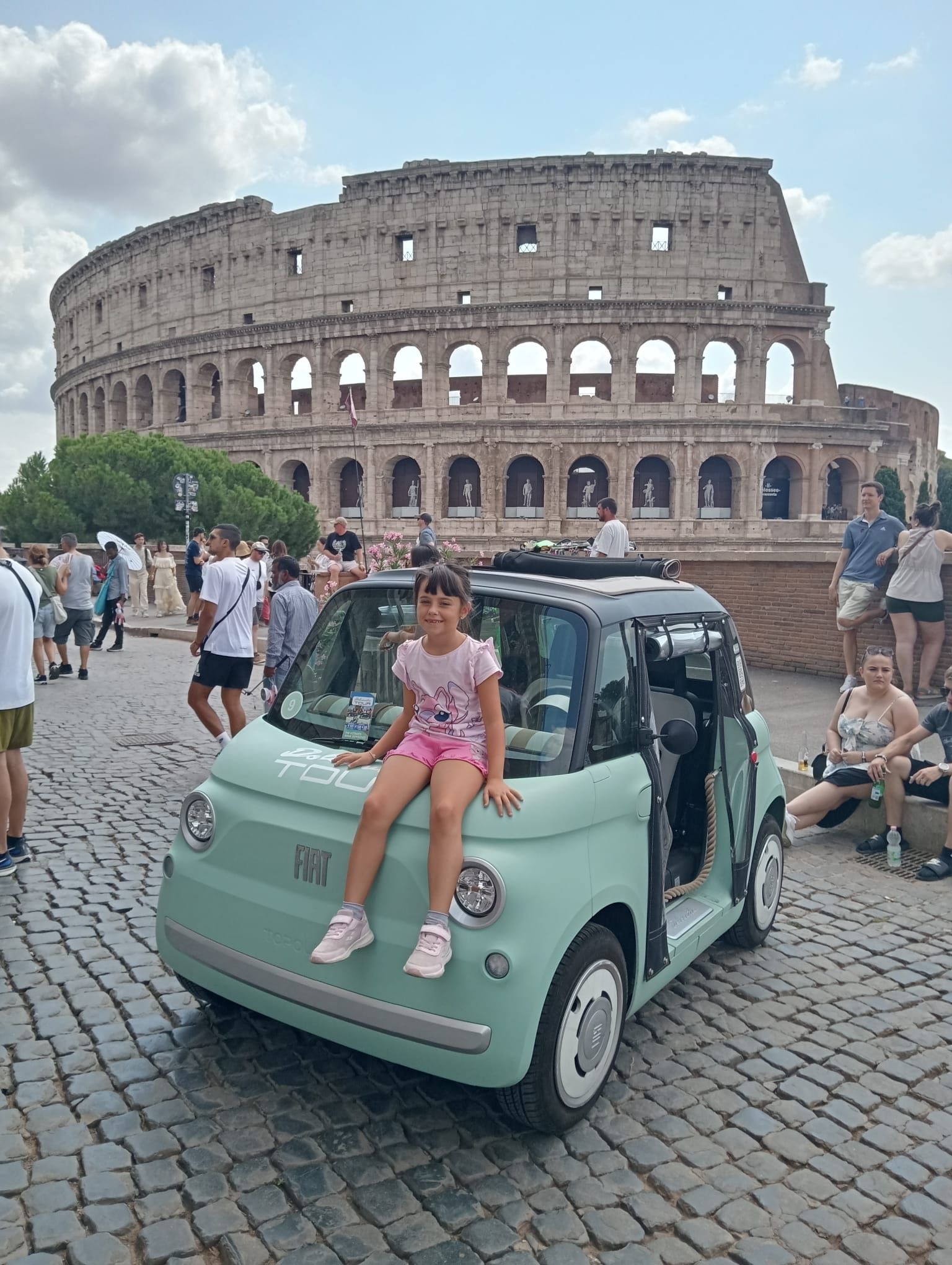 Mint microcar parked by the Colosseum with a girl sitting on the hood, tourists in the background