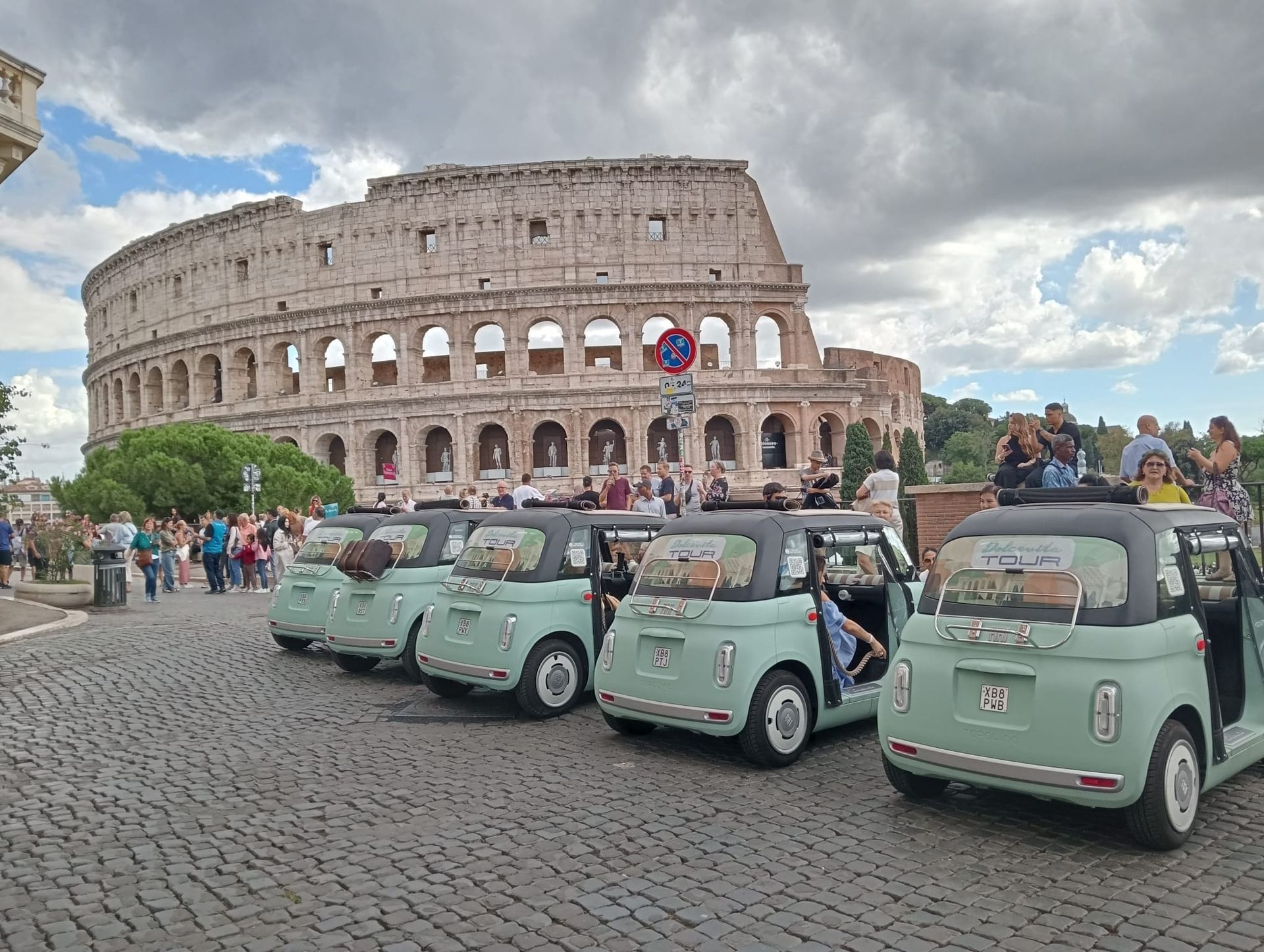 Vintage mint-green cars parked near the Colosseum in Rome, with tourists and cloudy sky.