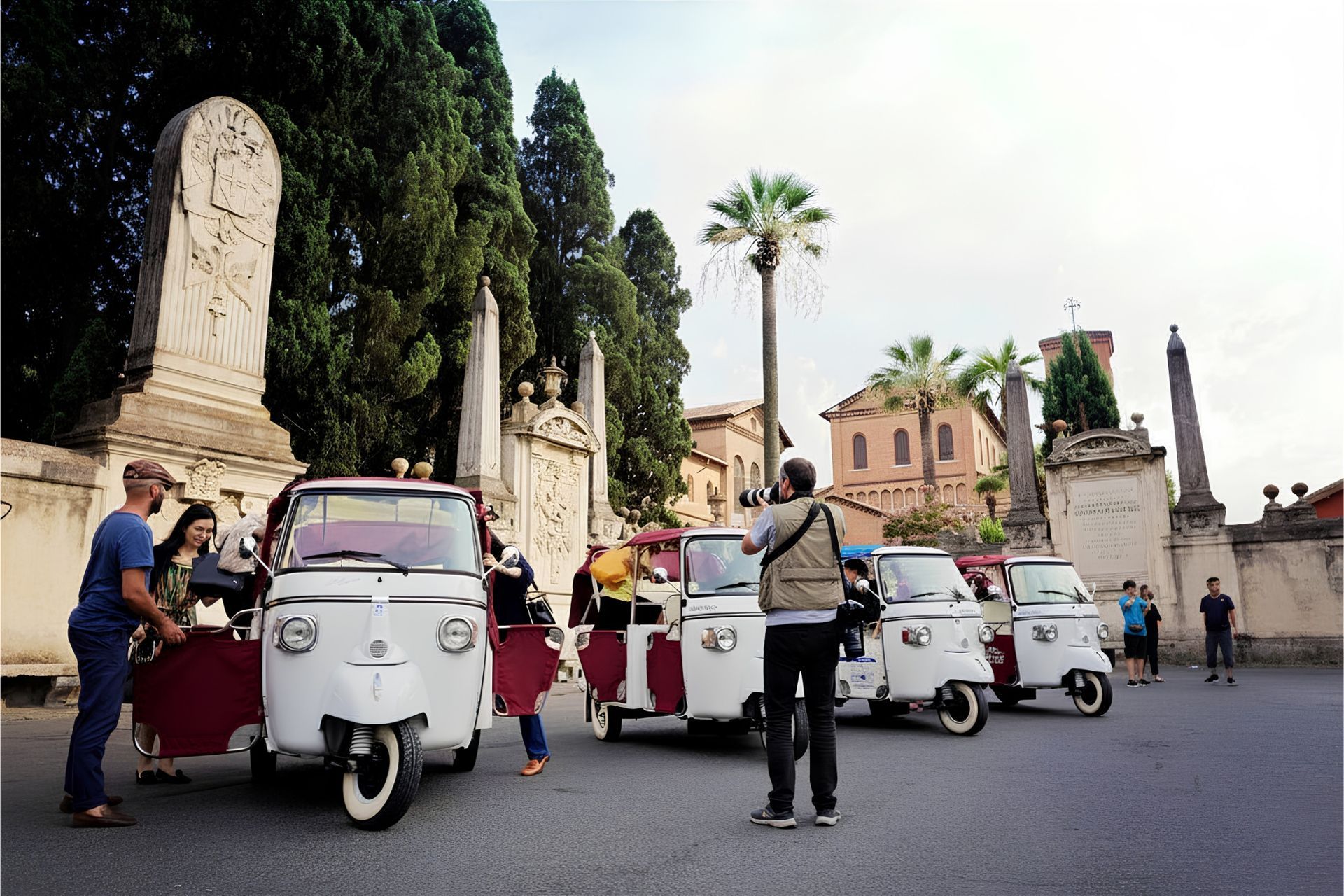 People posing with white tuk-tuks in a plaza near palm trees and stone monuments.