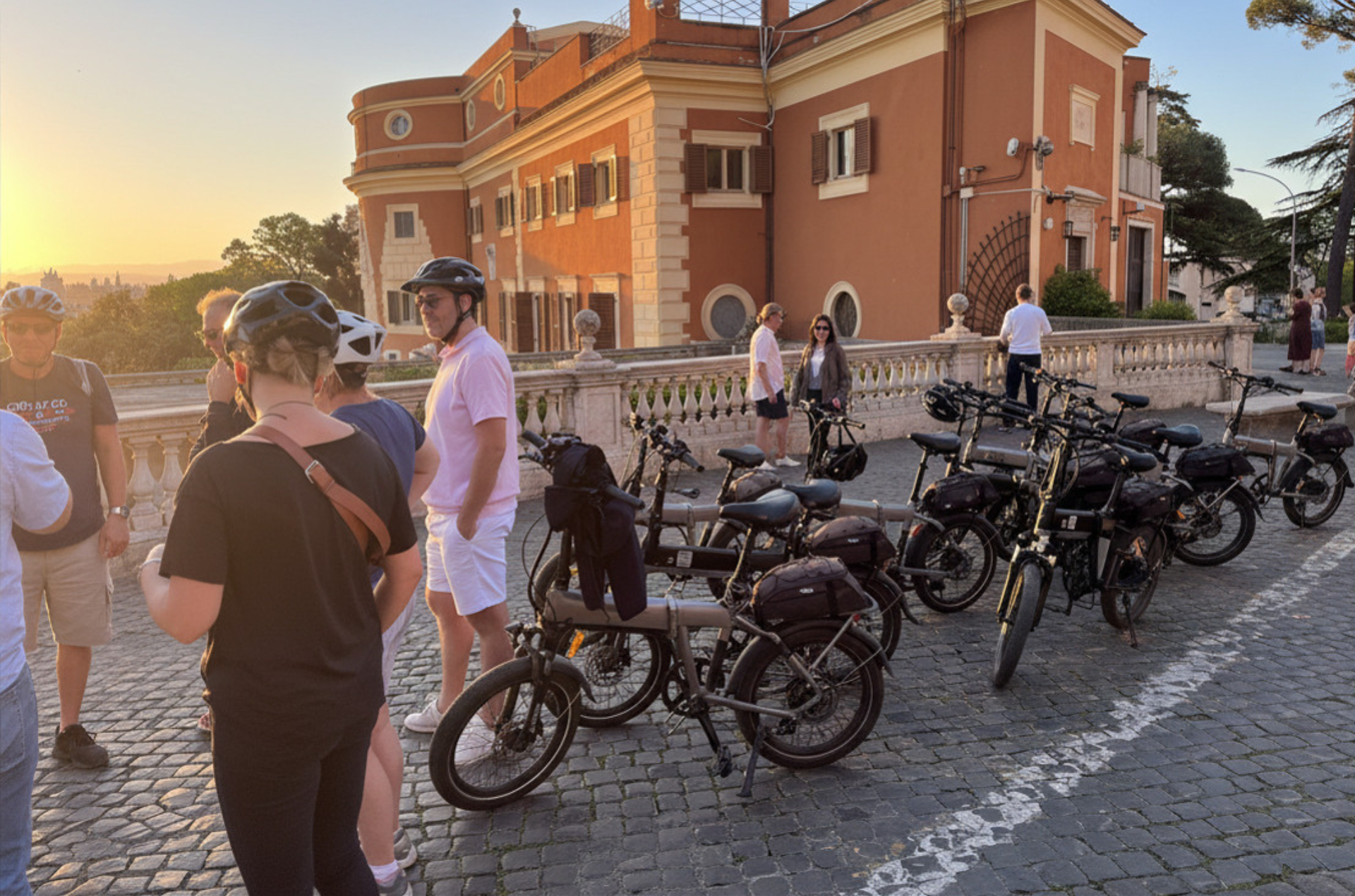 People standing beside parked motorcycles outside a red-brick building at sunset