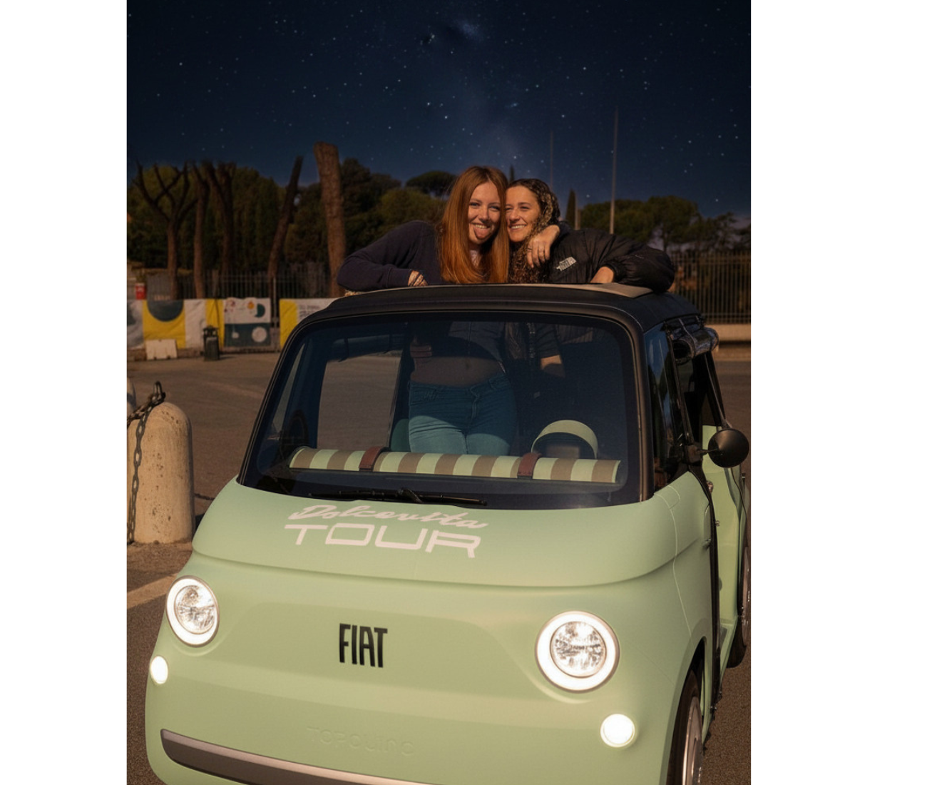 Two people leaning out of a mint-green Fiat 500 at night, smiling and posing.