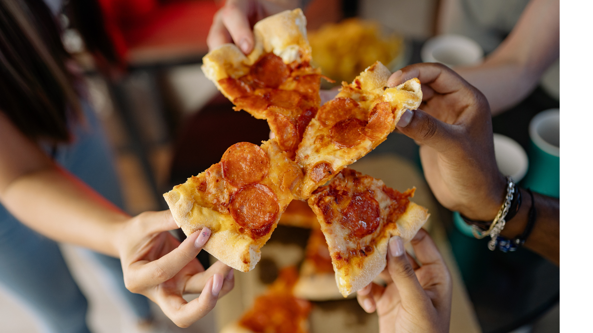 Hands sharing slices of pepperoni pizza over a table, with a blurred meal in the background