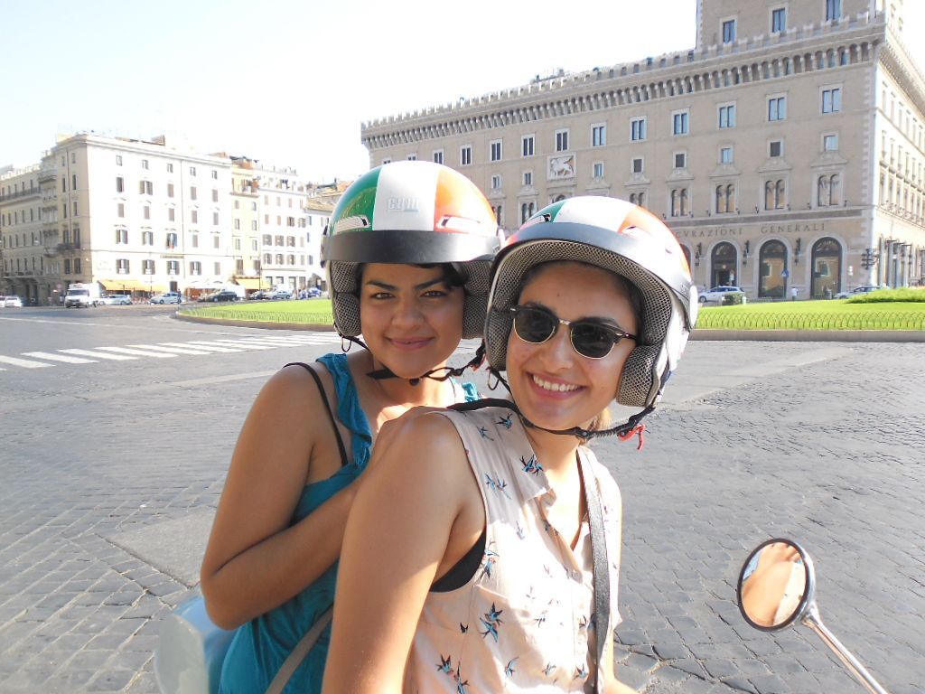 Two people in helmets smile on a scooter in a sunny city square.