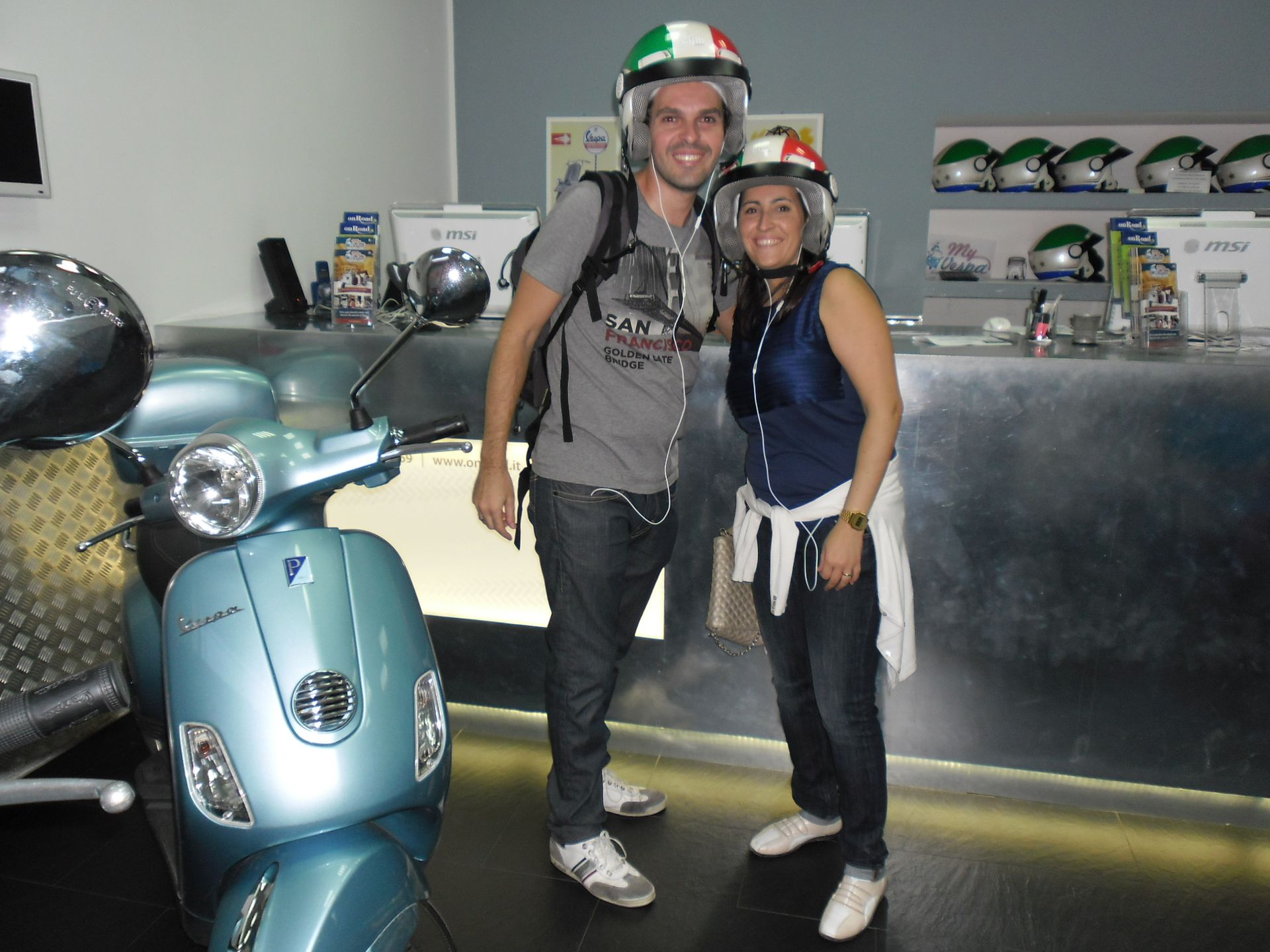 Two people standing beside a blue scooter in a helmet shop, smiling at the camera.