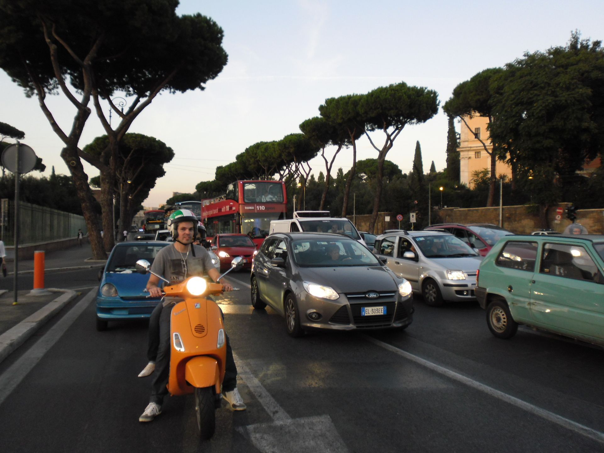 Traffic jam on a city street, with an orange scooter in front and cars lined up under tall trees.