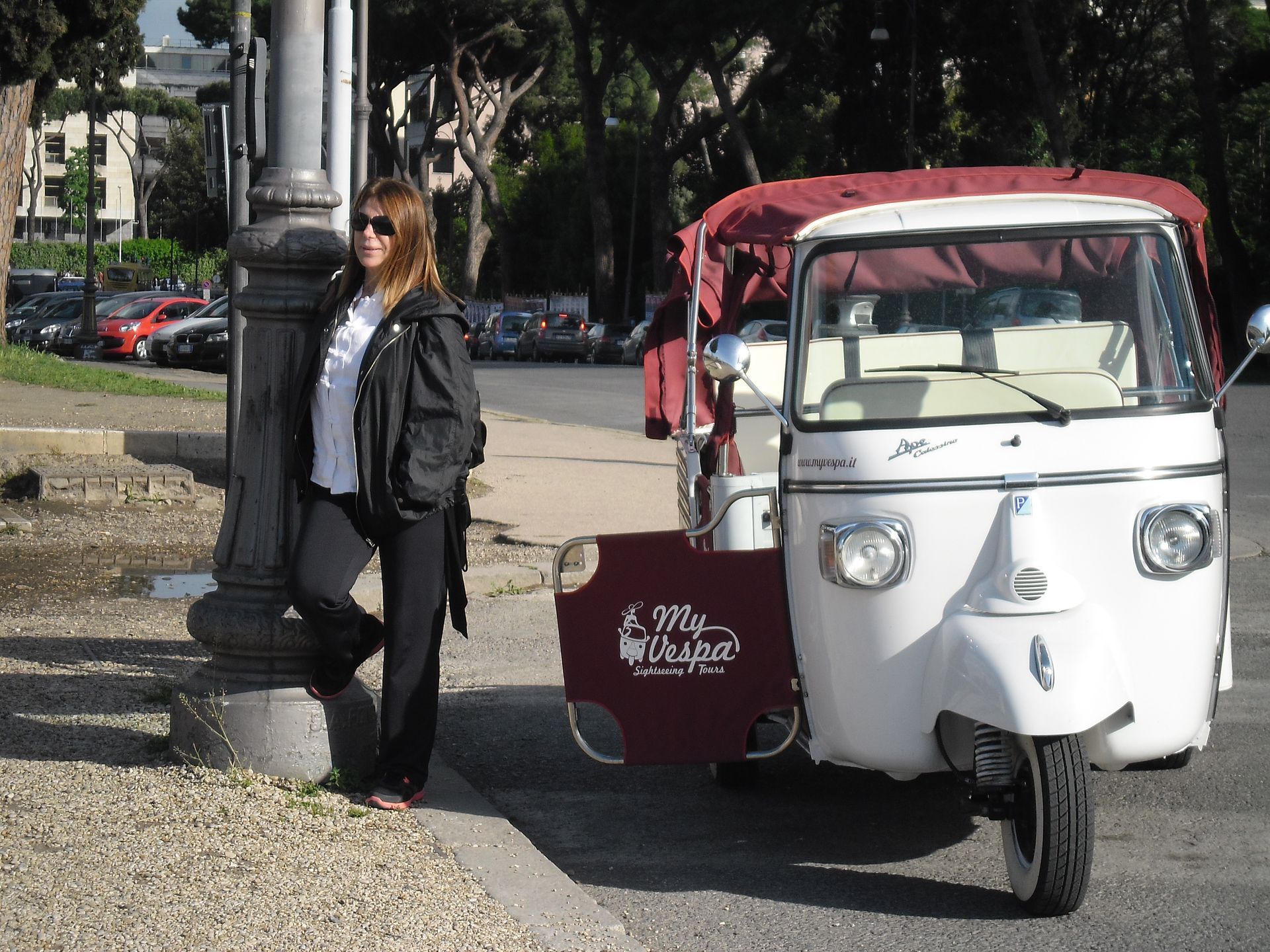 Woman standing beside a white-and-red tuk-tuk cart in a park area on a sunny day