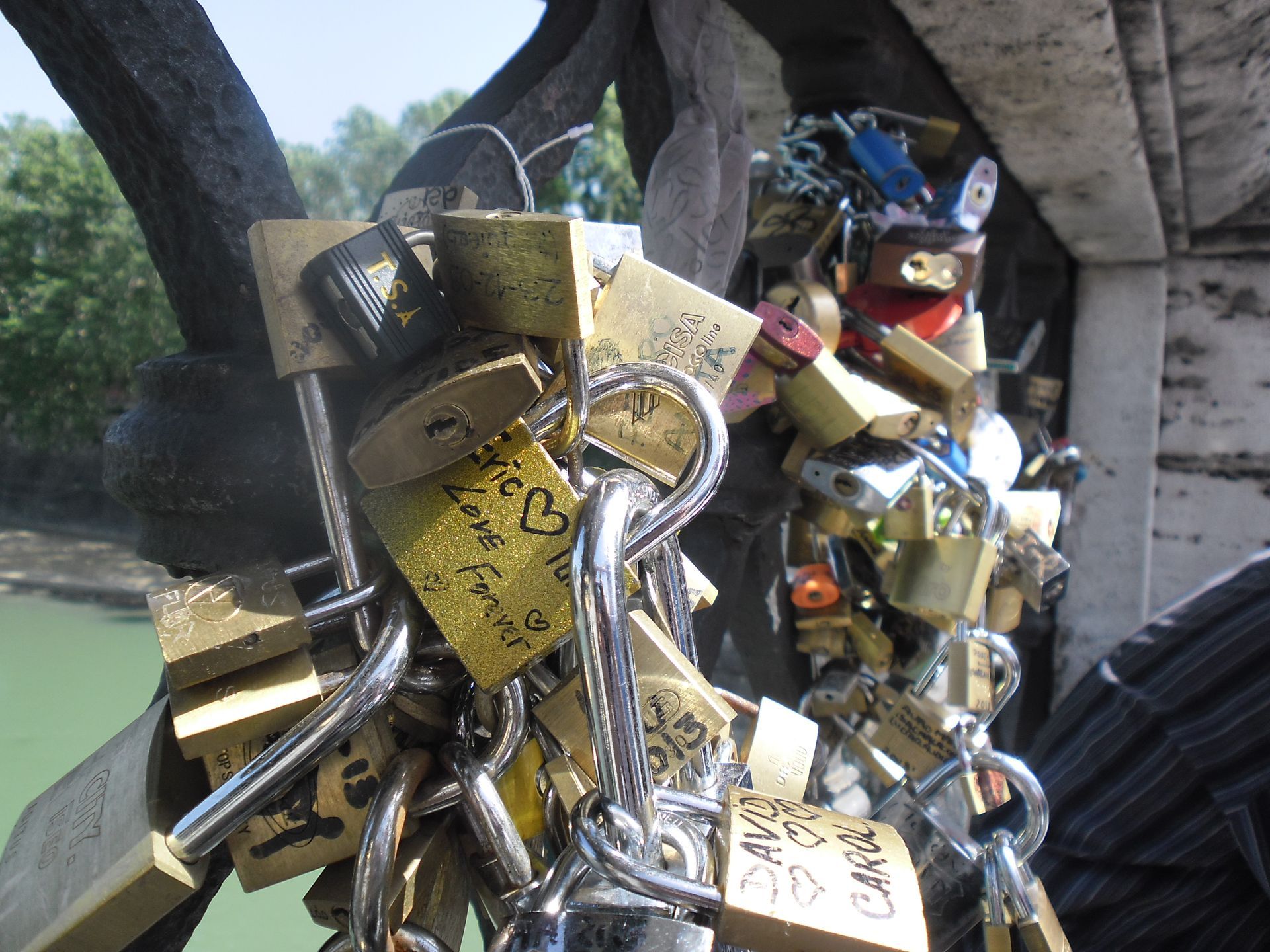 Cluster of gold padlocks on a metal fence near a river bridge