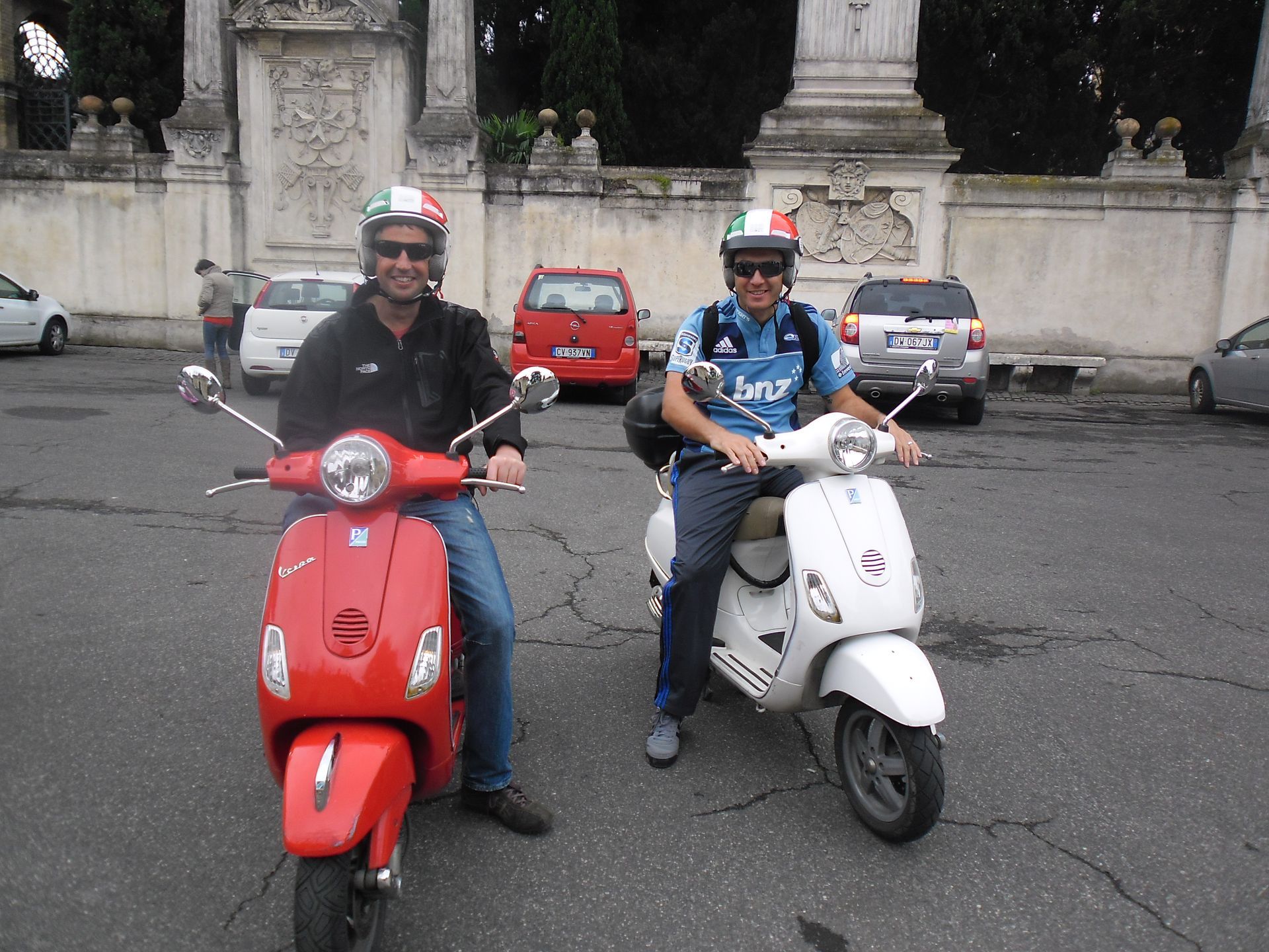 Two people on red and white scooters posing on a street in front of parked cars and a stone wall.