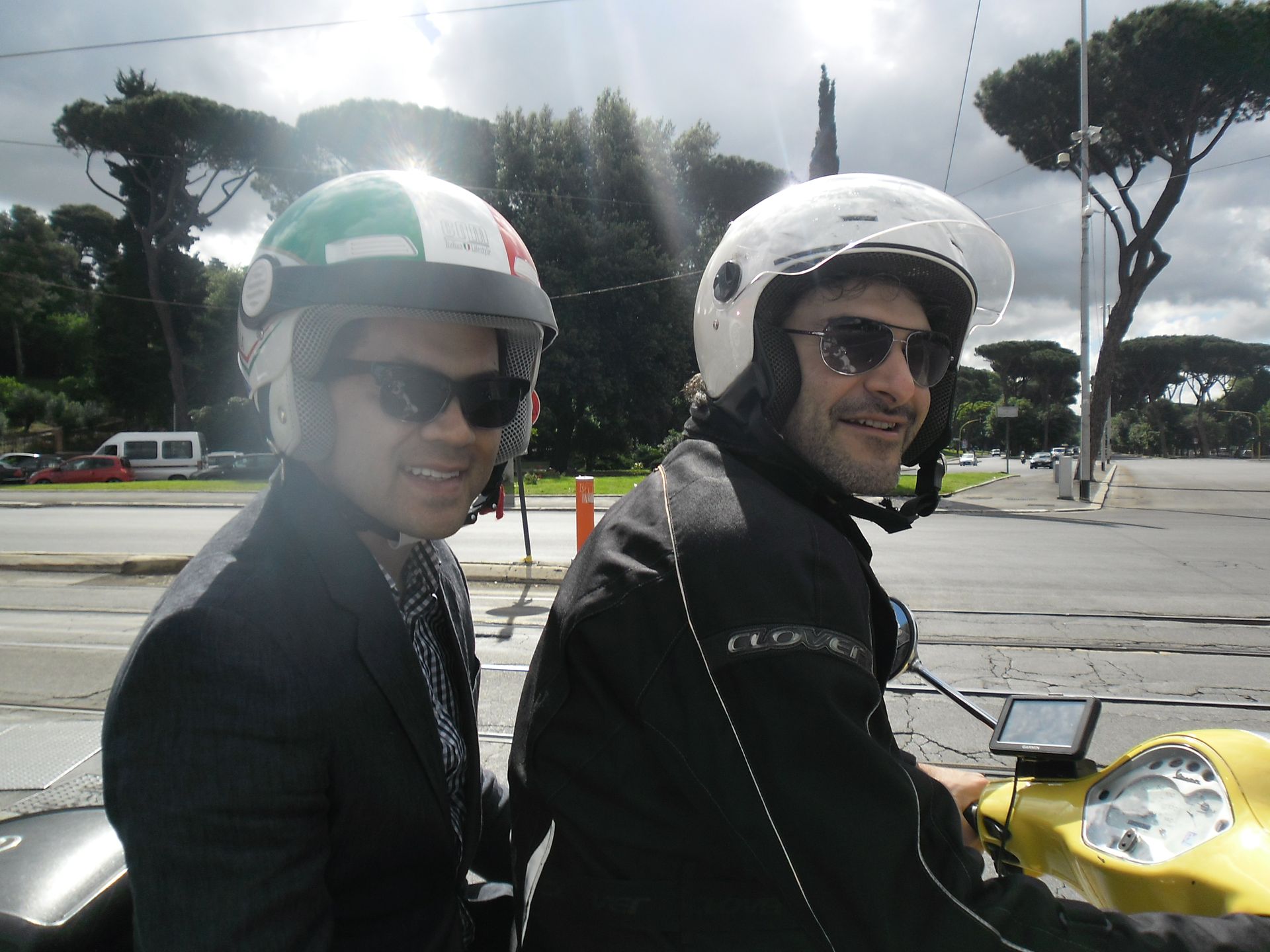 Two people on a yellow scooter wearing helmets and sunglasses on a sunny street.