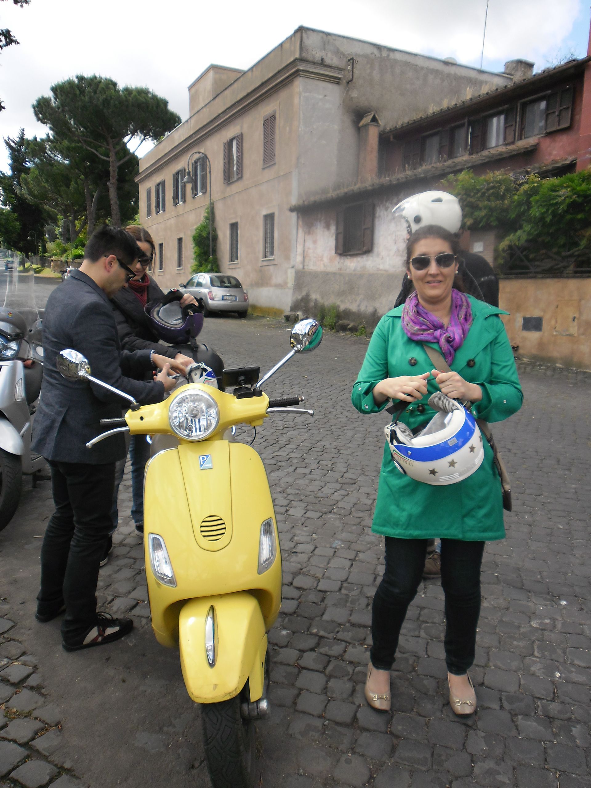 Woman in green coat holding a helmet beside a yellow scooter on a city street, with a man behind her.