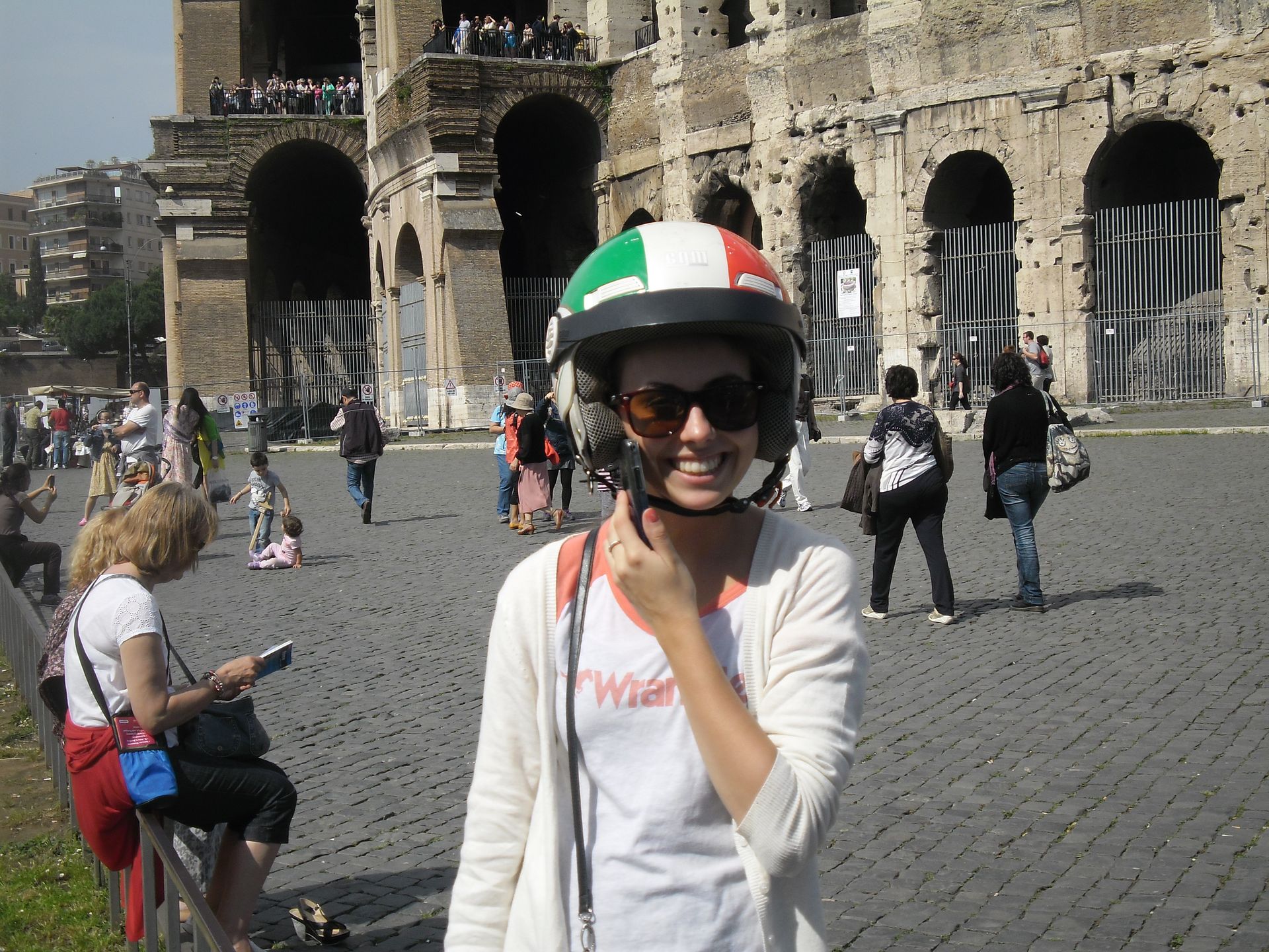 Person in an Italian flag helmet smiles in front of a historic stone building and archway.