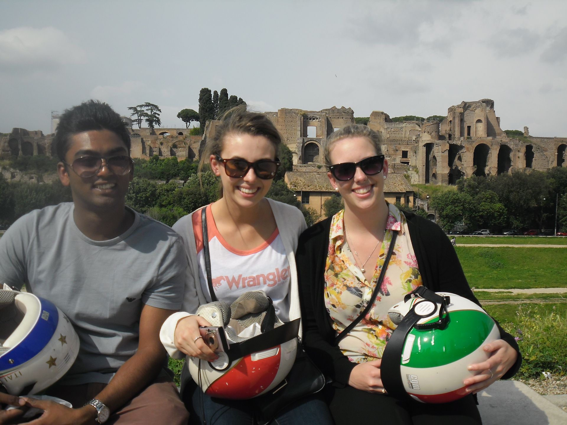Three people sitting outdoors with helmets, in front of ancient stone ruins on a sunny day
