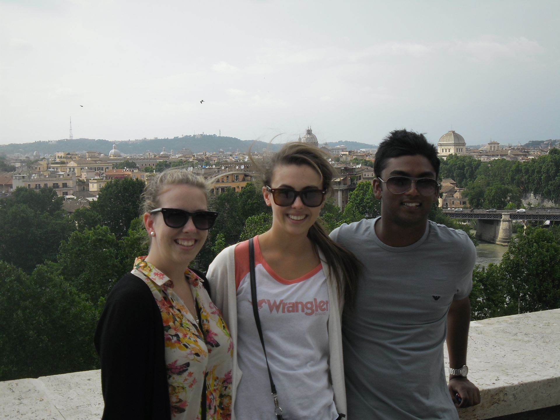 Three smiling people standing by a stone wall with a cityscape and cloudy sky in the background