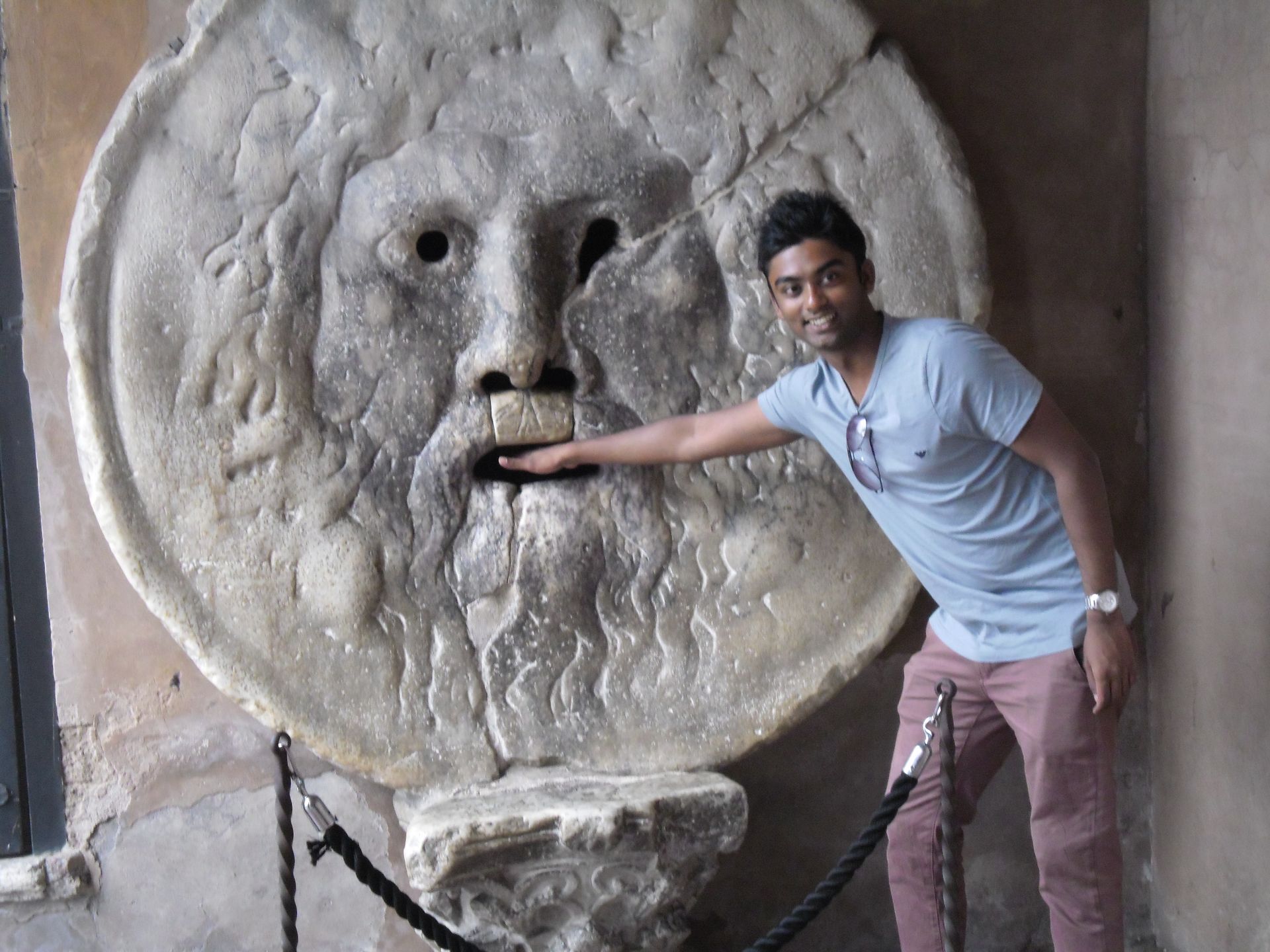 Man posing beside a giant stone face sculpture with a hand in its mouth
