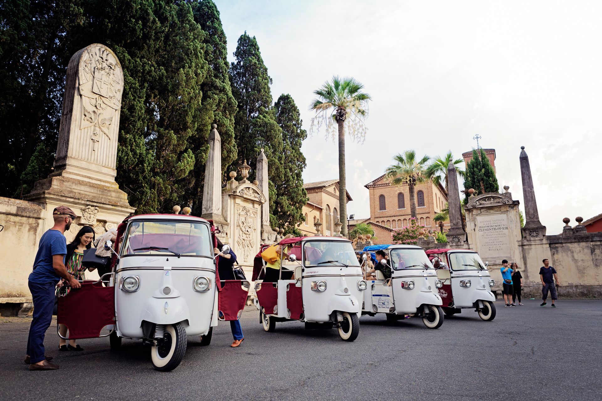 White tuk-tuks parked in a historic square with people and tall cypress trees behind them.