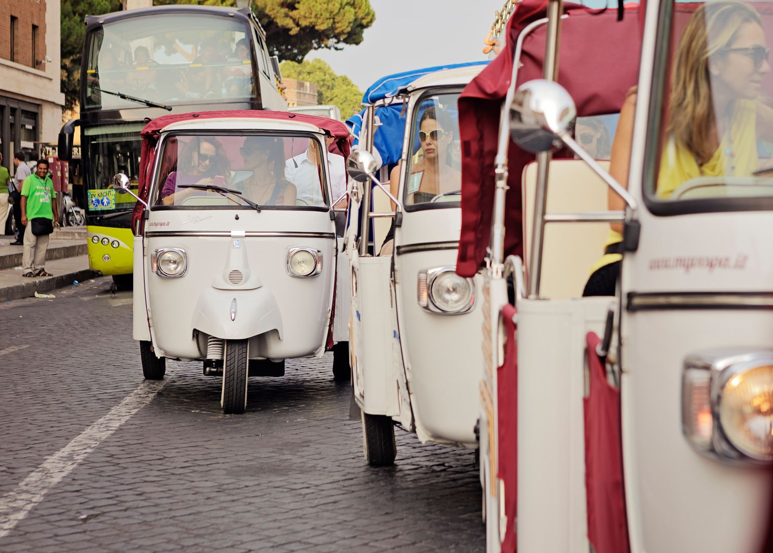 White tuk-tuks line a cobblestone street in traffic, with pedestrians and a bus in the background.