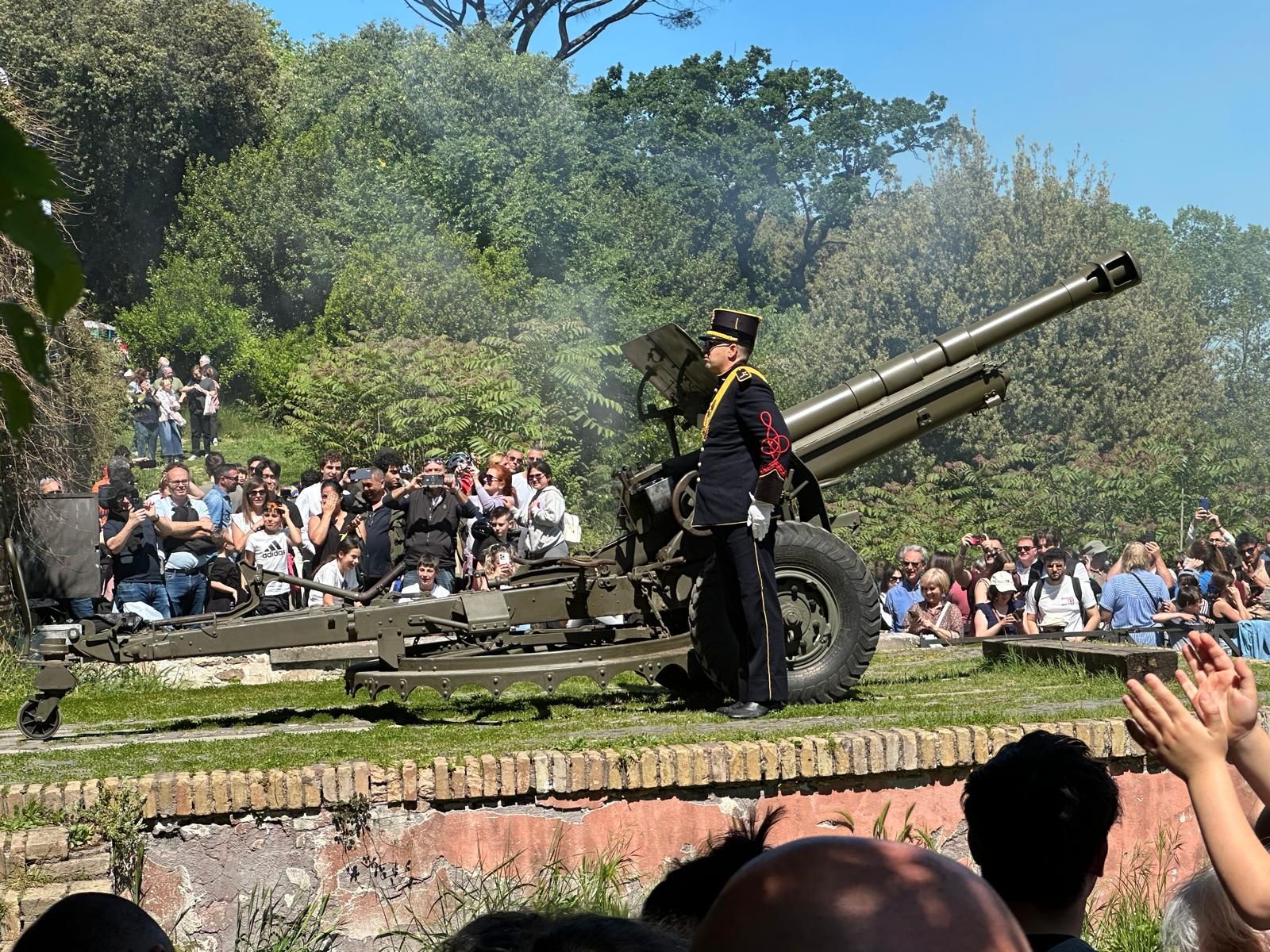 Cannon salute at an outdoor crowd event, with a uniformed guard beside the artillery piece.