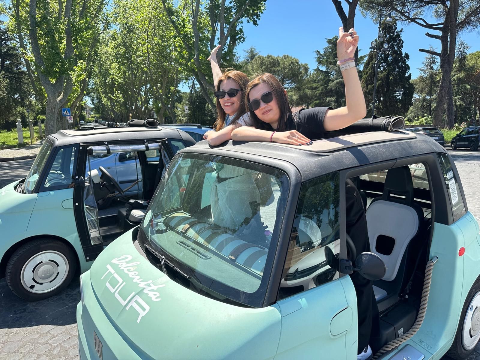 Two people leaning from a turquoise car with roof open, waving in a sunny outdoor setting