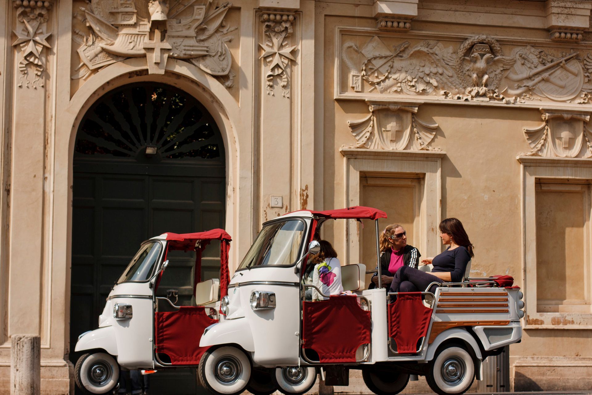 White and red tourist tuk-tuks parked beside an ornate stone building, with seated passengers.