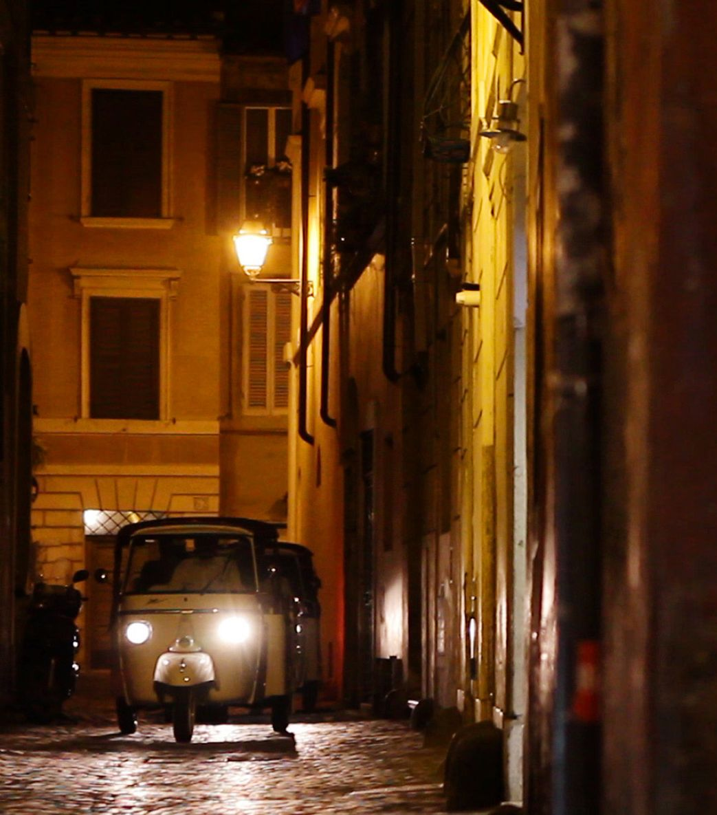 Small white car with headlights on in a narrow, dimly lit cobblestone street at night