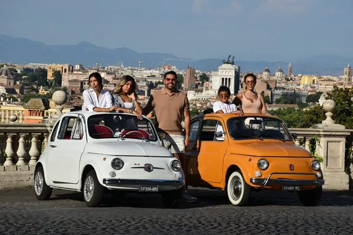 Two vintage Fiat 500s parked with people on a scenic city overlook with a skyline in the background