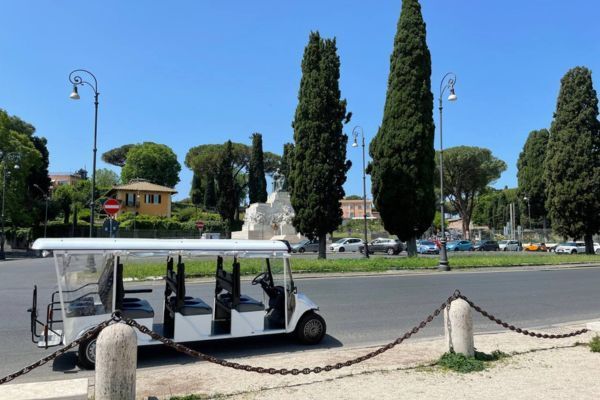 Open-sided tourist tram parked by a road with tall cypress trees and a sunny plaza in the background