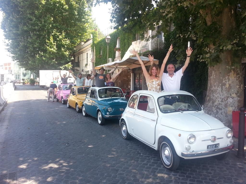 Line of vintage cars on a cobblestone street, with people cheering from the lead white car.