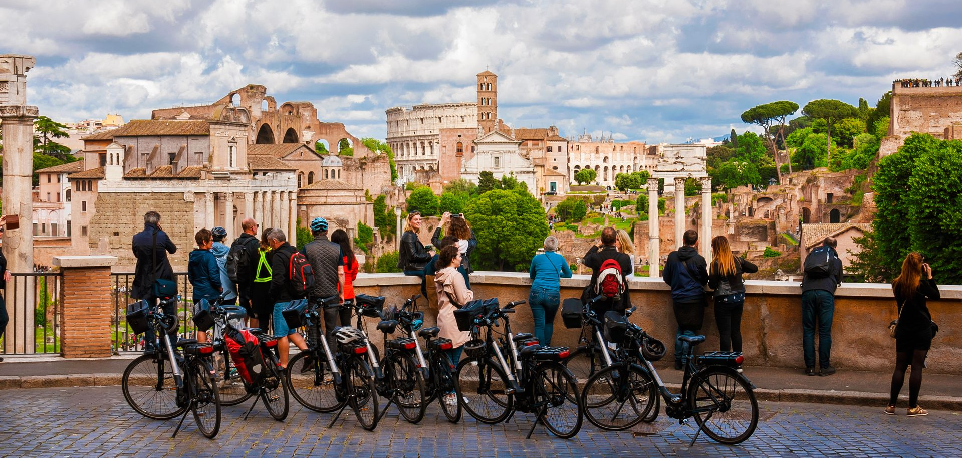 Cyclists pause on a bridge overlooking a historic cityscape under cloudy skies.