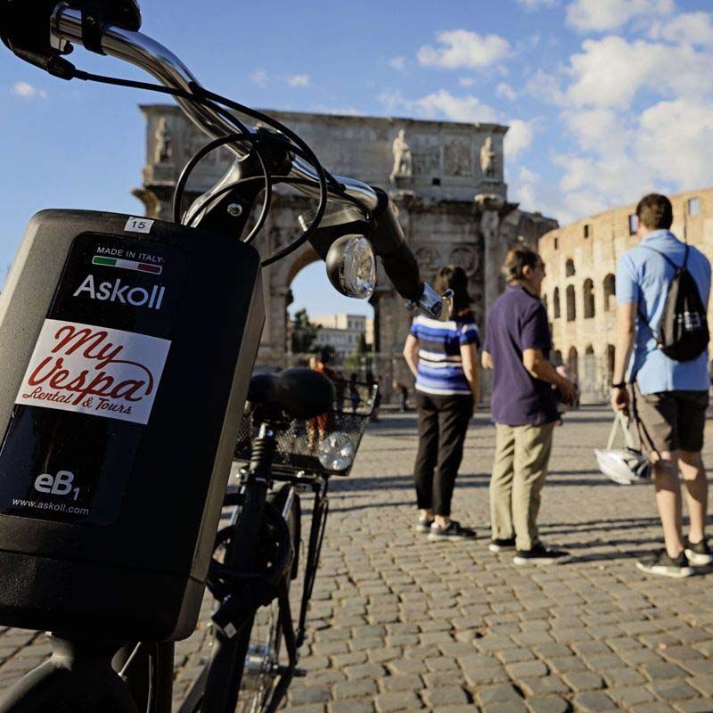 Bicycle handlebars in foreground; people stand on a cobblestone street near the Colosseum in Rome.