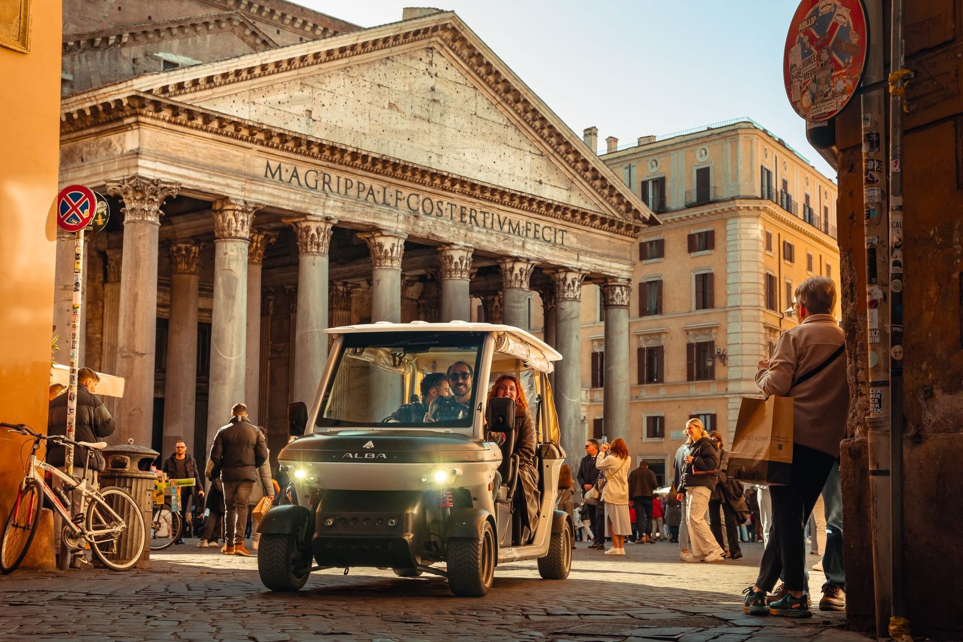 Small electric shuttle in a sunlit cobblestone square outside a grand classical building, with pedestrians nearby