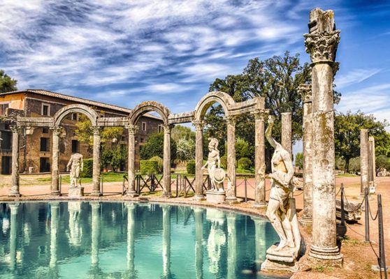 Ancient ruins with arched columns beside a turquoise pool under a cloudy sky