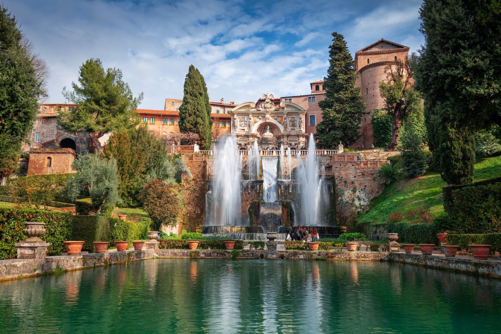 Terraced garden with twin fountains, lush greenery, and historic buildings beside a turquoise pool