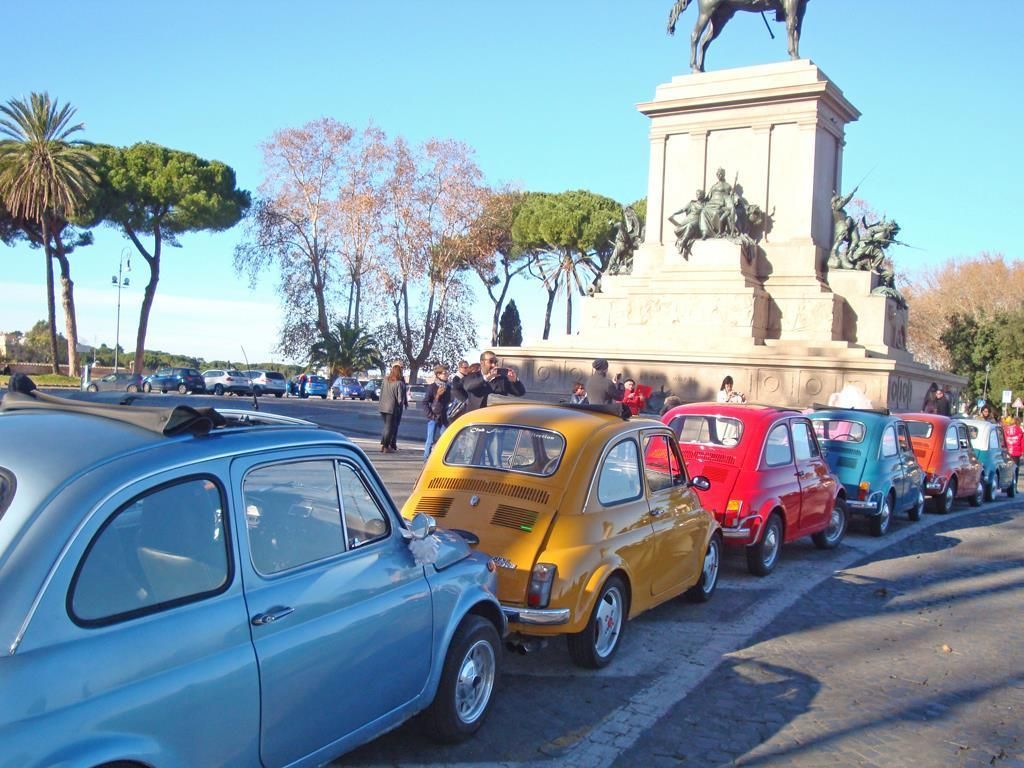 Line of colorful vintage cars parked beside a plaza statue on a sunny day