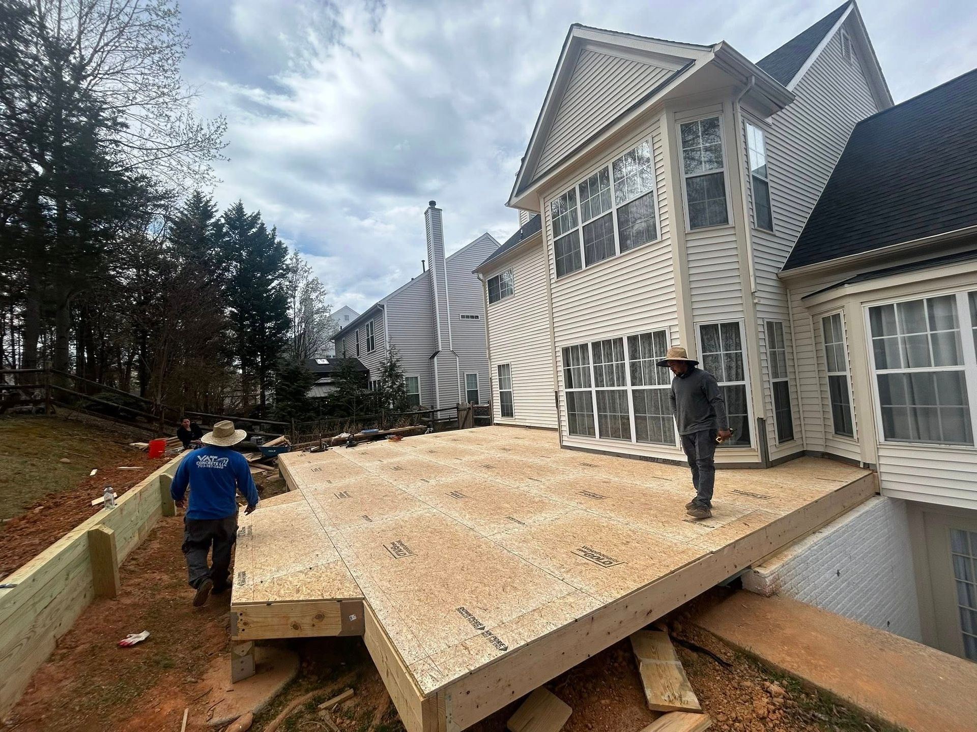 Construction workers building a wooden deck attached to a beige two-story house. Cloudy day.