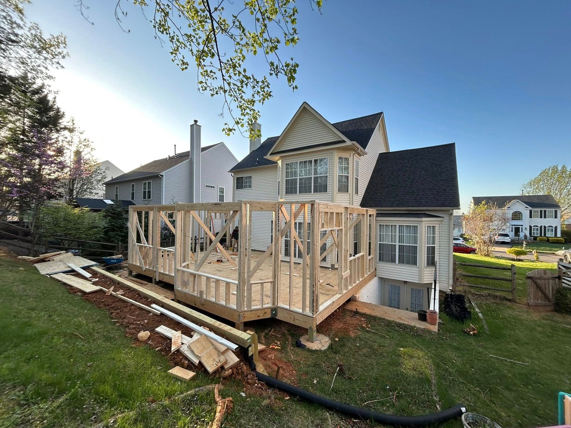 Wooden deck under construction attached to a two-story house, on a grassy hill.