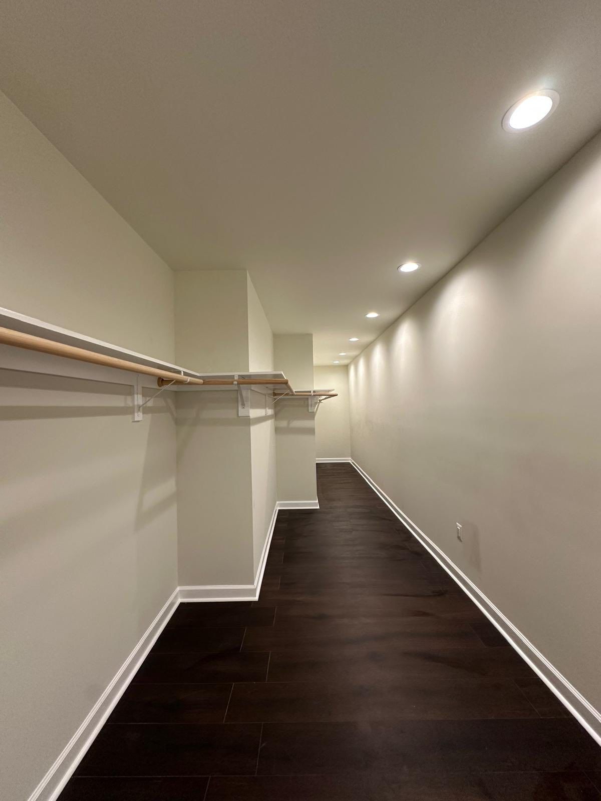 Narrow walk-in closet with dark wood floors, white walls, and overhead lighting; contains a clothes rod and shelving.