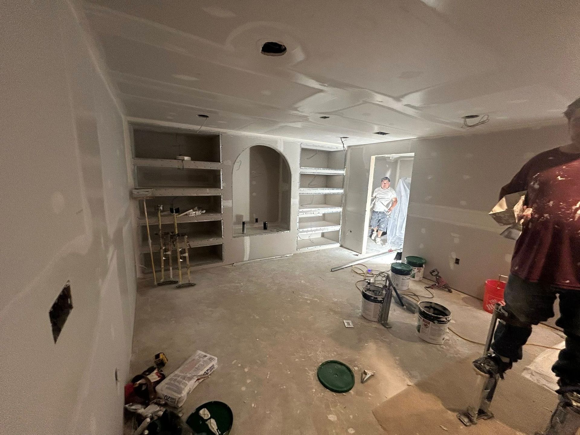 Room under renovation with drywall, shelves, and a worker on stilts. Another person stands in doorway.