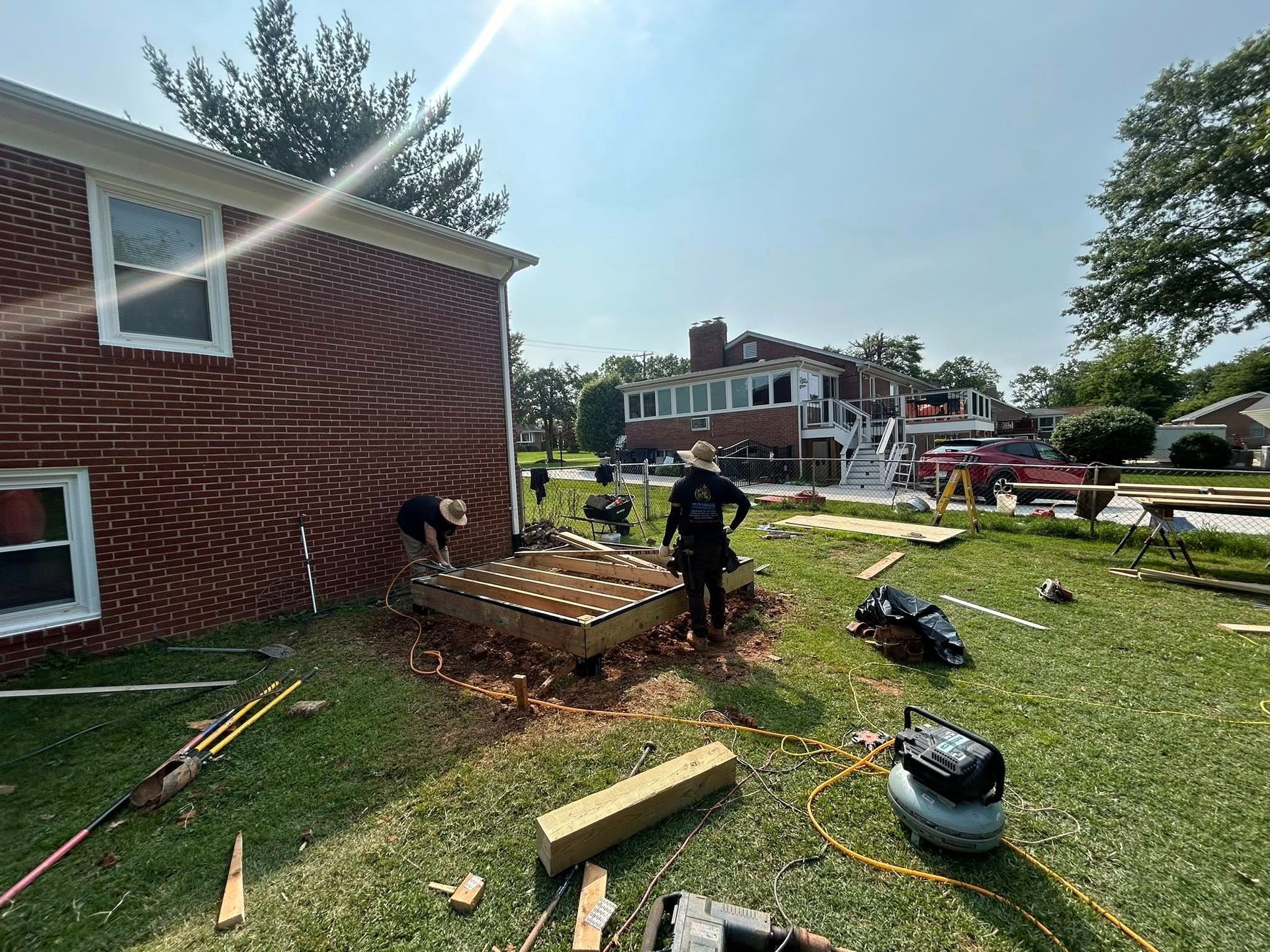Two construction workers building a deck next to a red brick house on a sunny day.