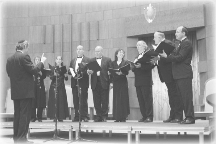 Choir of formally dressed singers performing on a stage, holding music folders and standing behind microphones.