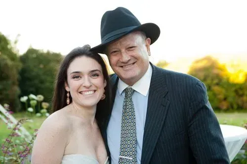 Smiling couple at an outdoor event, the man in a dark suit and hat beside a woman in a strapless dress.