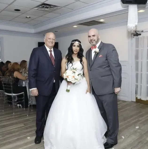 Bride in white gown stands between two men in suits at an indoor wedding reception.