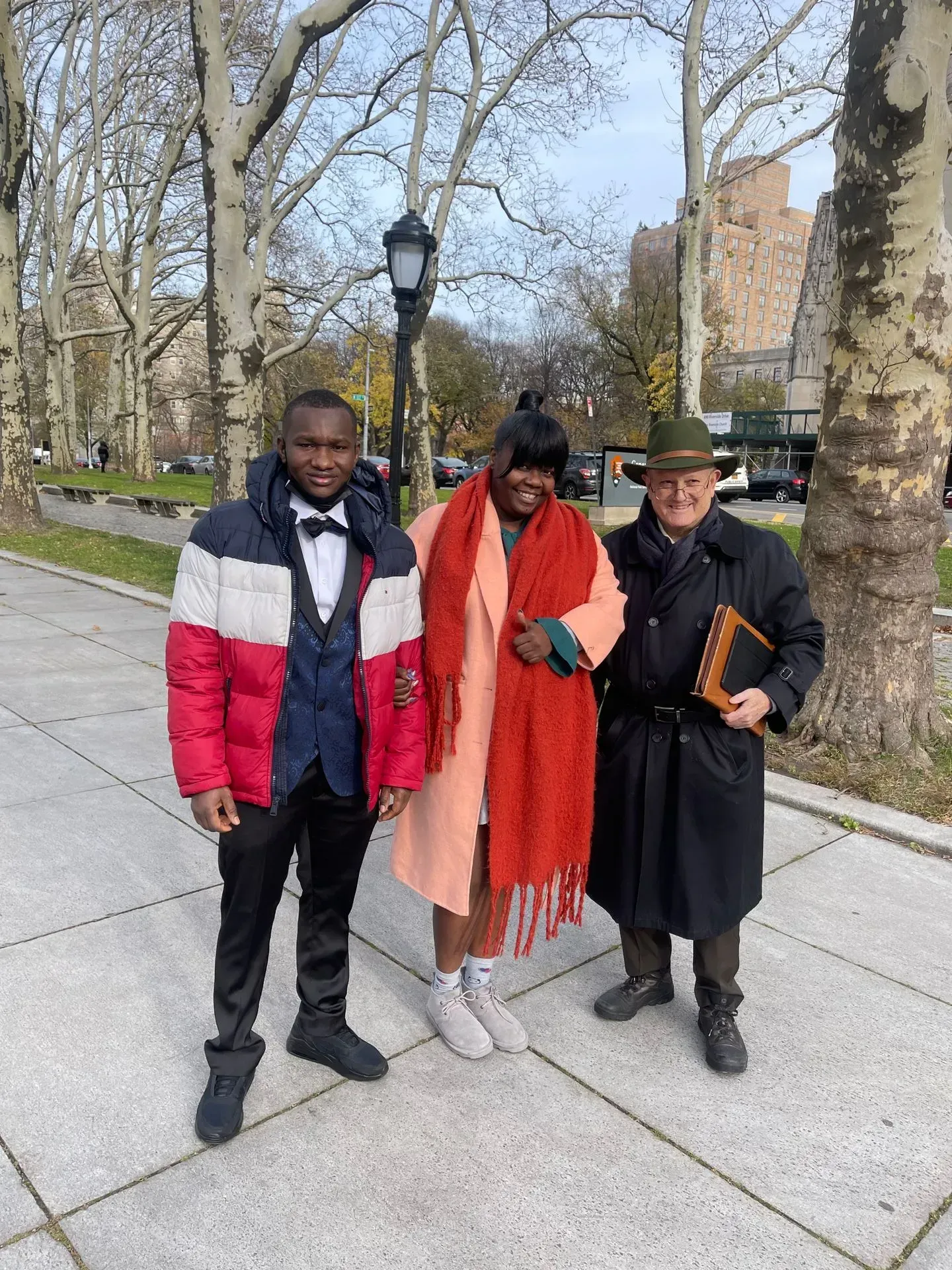 Three people standing on a tree-lined sidewalk, wearing winter coats and scarves, posing outdoors.